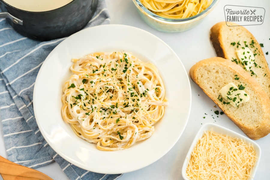 A bowl of Fettuccine Alfredo with homemade Alfredo sauce next to sliced bread and parmesan cheese.