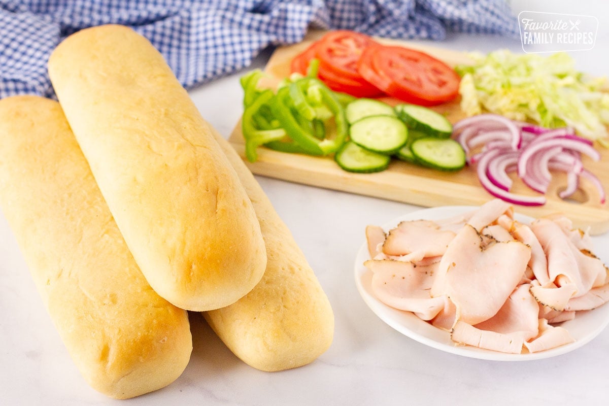 Three loaves of baked Subway bread with a plate of meat and cutting board of vegetables for a sandwich.