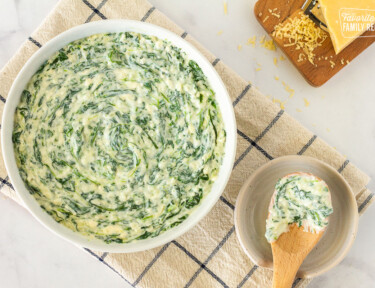 Top view of a bowl of Creamed Spinach with a wooden serving spoon on the side on a saucer. Board of fresh grated Parmesan cheese on the side.