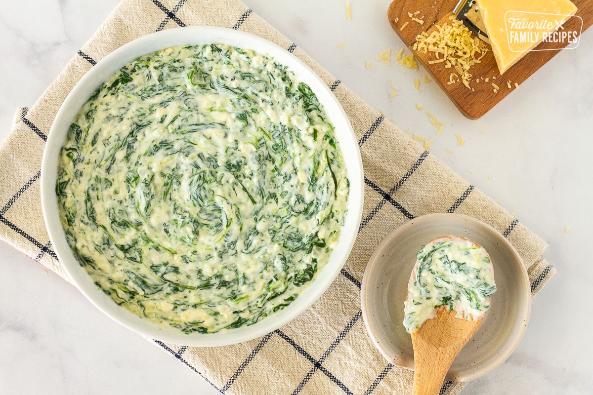 Top view of a bowl of Creamed Spinach with a wooden serving spoon on the side on a saucer. Board of fresh grated Parmesan cheese on the side.