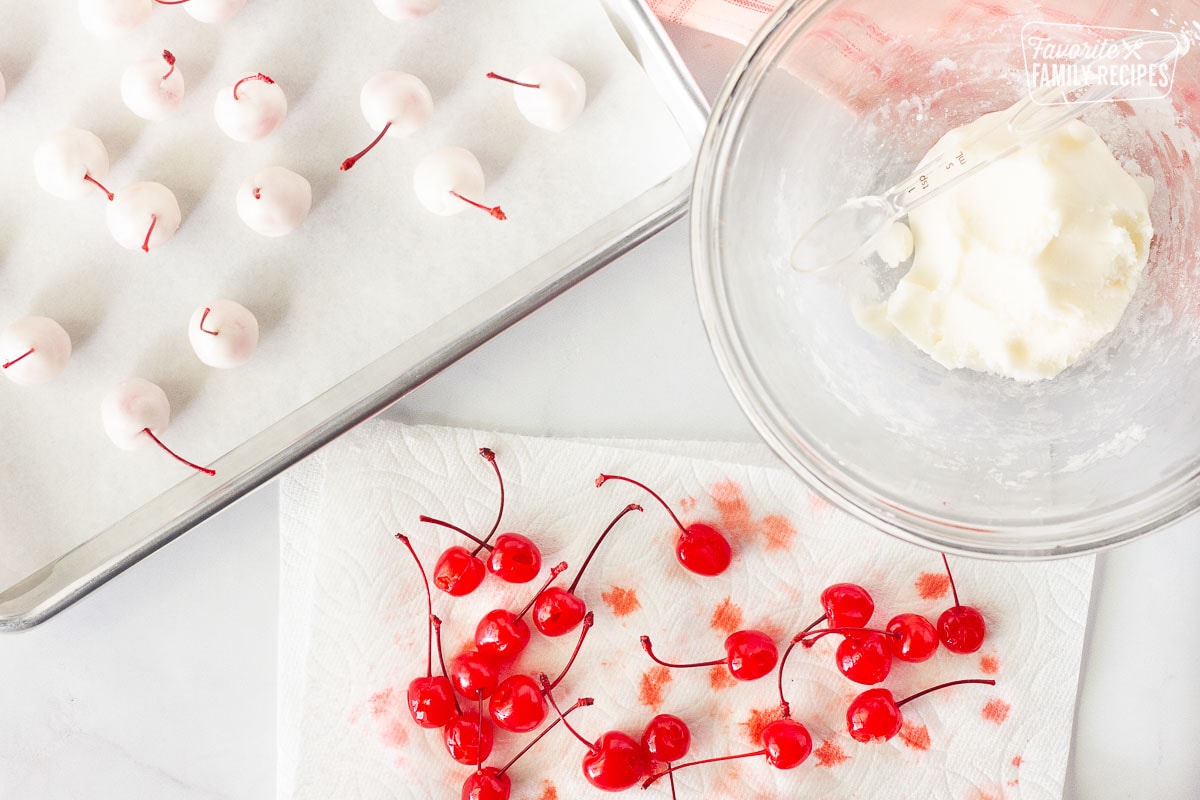 Baking sheet with fondant covered cherries, a bowl of fondant with measuring spoon and a paper towel with cherries for Chocolate Covered Cherries.