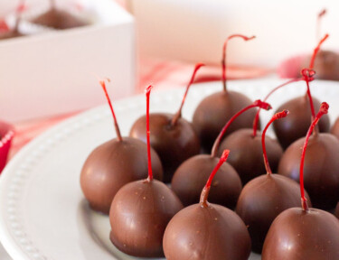 Close up plate of Chocolate Covered Cherries with gift boxes of cherries behind.