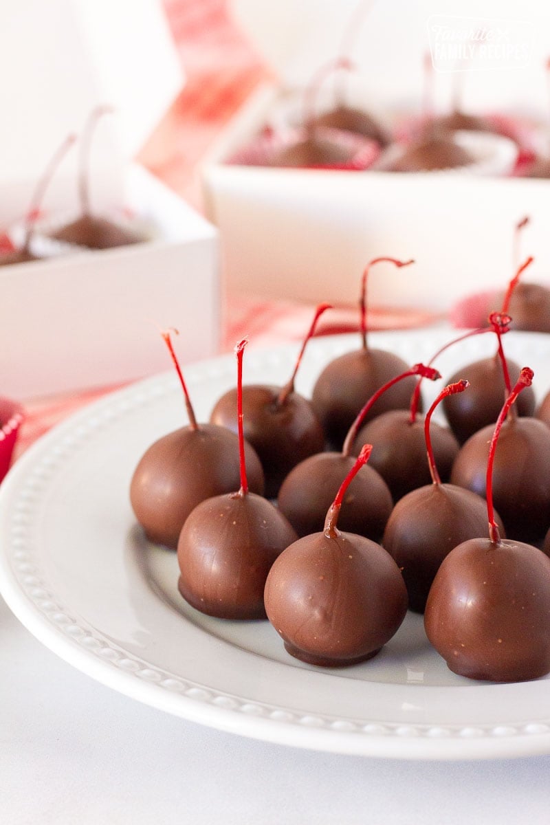 Close up plate of Chocolate Covered Cherries with gift boxes of cherries behind.