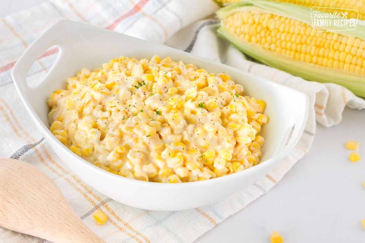 Bowl of Creamed Corn with a wooden spoon and fresh corn on the side.