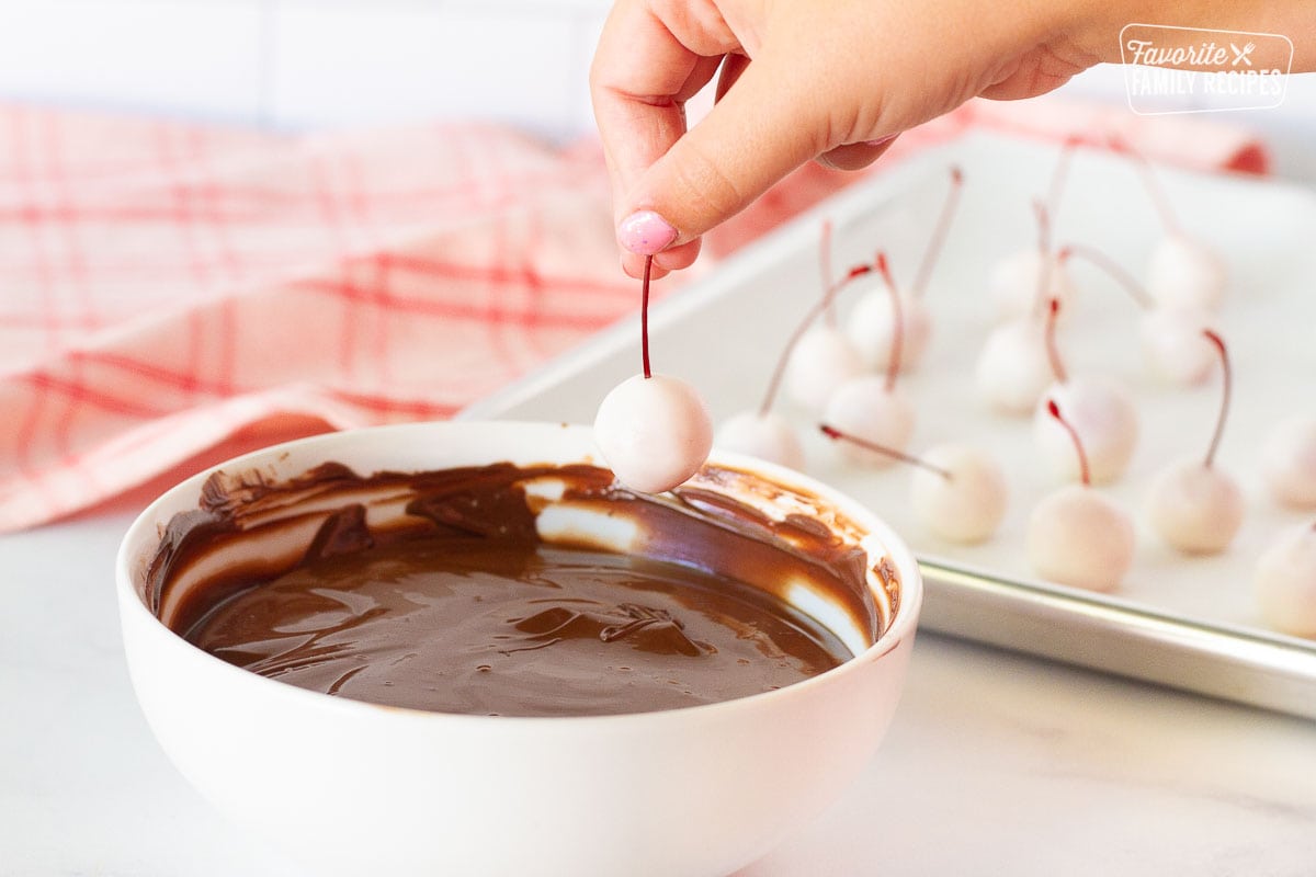 Hand dipping fondant cherry into a bowl of melted chocolate for Chocolate Covered Cherries.