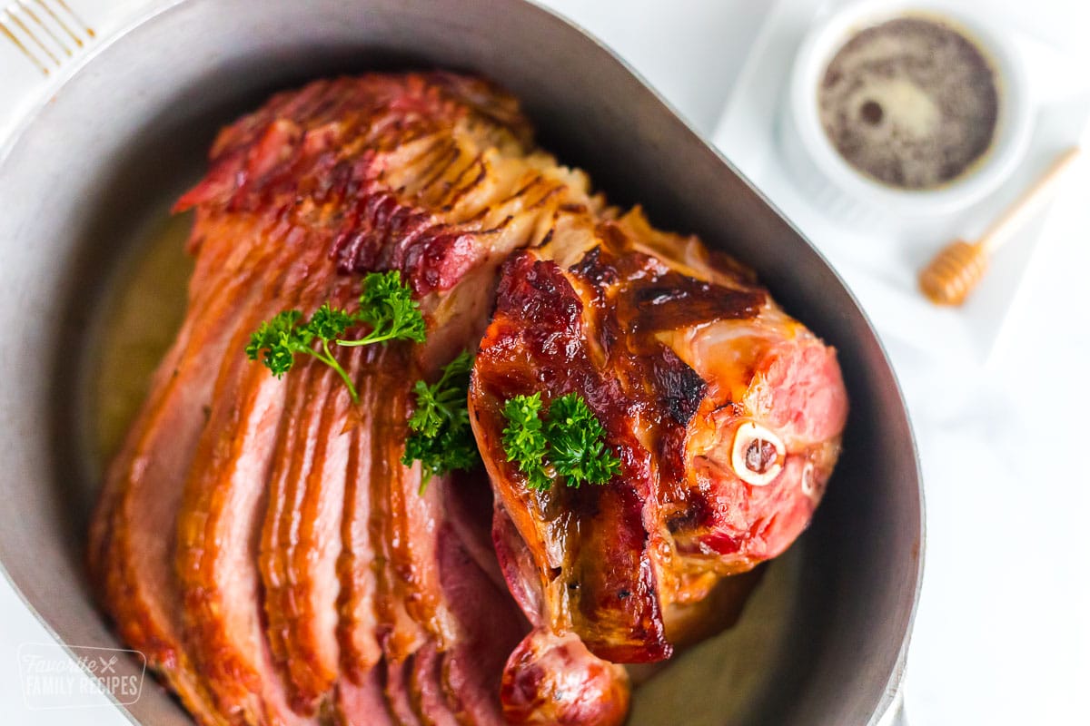 A whole honey baked ham in a roasting pan next to a bowl of honey