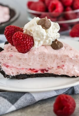 A slice of Raspberry Cream Pie on a plate topped with raspberries, whipped cream, and chocolate chips