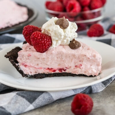 A slice of Raspberry Cream Pie on a plate topped with raspberries, whipped cream, and chocolate chips