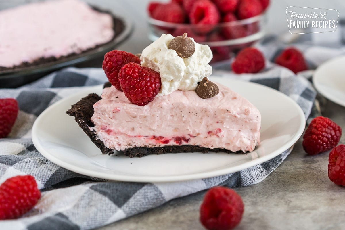 A slice of Raspberry Cream Pie on a plate topped with raspberries, whipped cream, and chocolate chips
