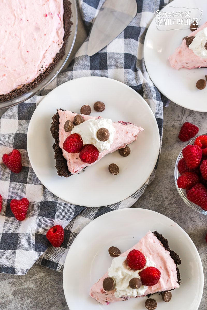 Three slices of Raspberry Cream Pie on plates topped with whipped cream, raspberries, and chocolate chips
