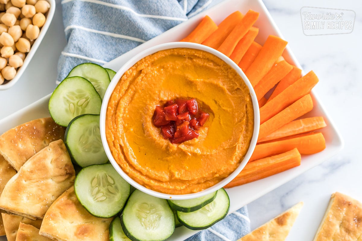 A tray with a bowl of roasted red pepper hummus, carrot sticks, cucumber slices, and pita chips
