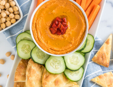 A tray with a bowl of roasted red pepper hummus, carrot sticks, cucumber slices, and pita chips