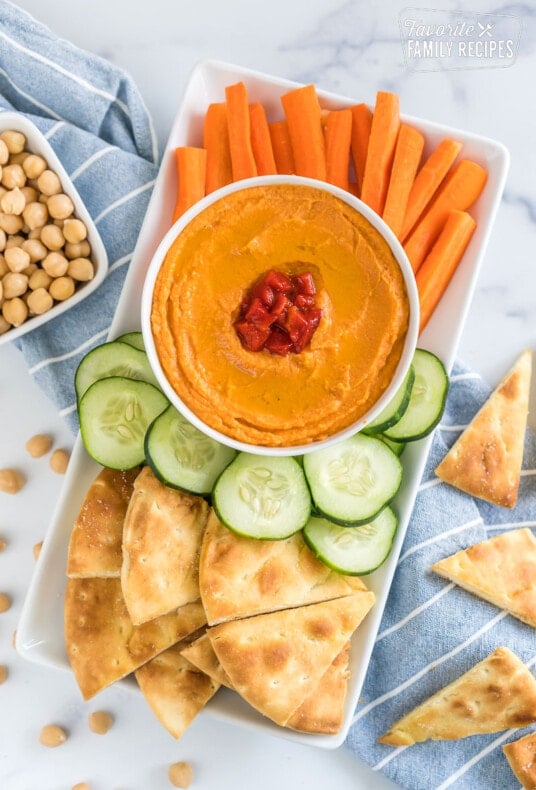 A tray with a bowl of roasted red pepper hummus, carrot sticks, cucumber slices, and pita chips