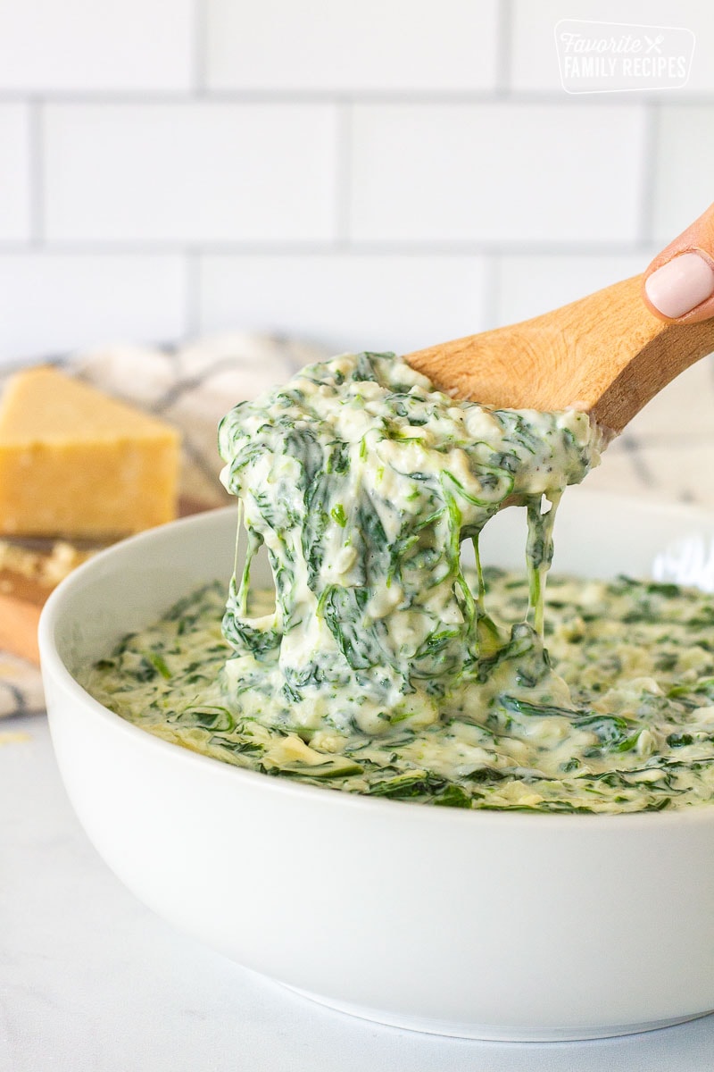 Hand pulling up a scoop of Creamed Spinach on a wooden spoon.