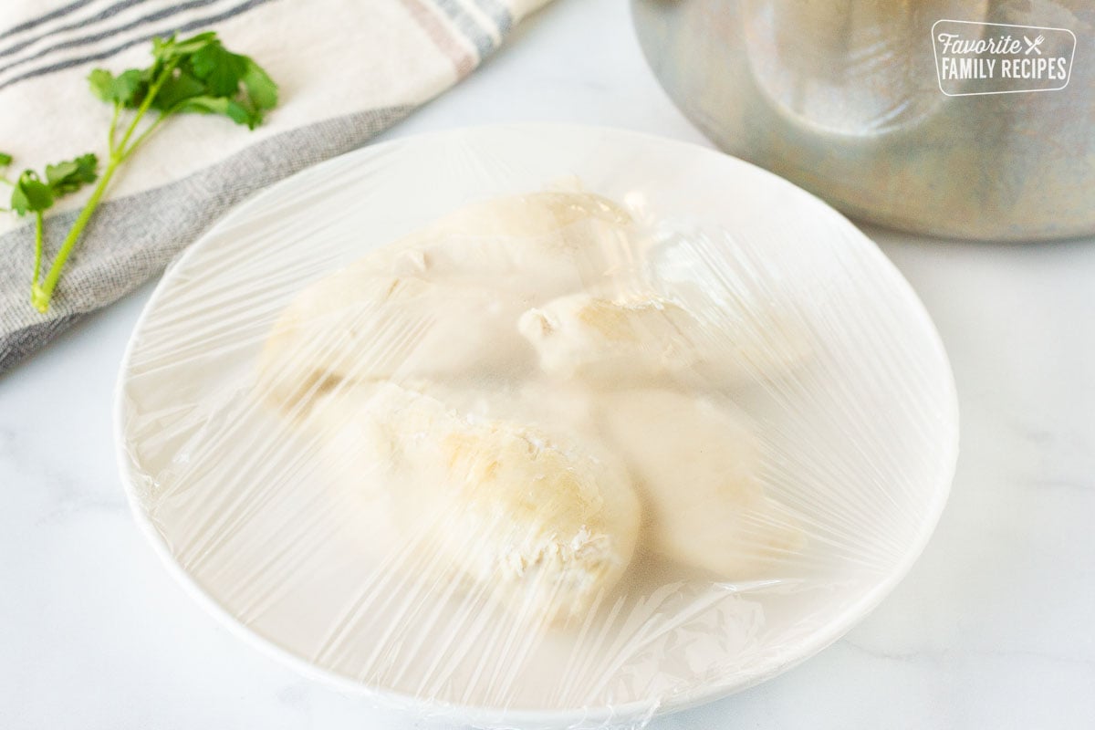 Plastic wrap over a plate of cooked chicken for Homemade Chicken Noodle Soup.