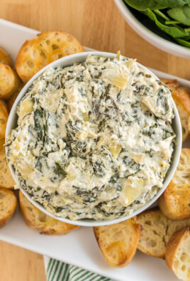 A bowl of crockpot spinach artichoke dip surrounded by slices of baguette