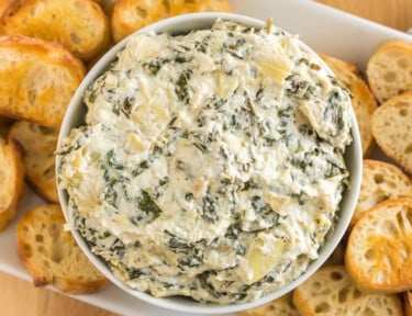 A bowl of crock pot spinach artichoke dip surrounded by slices of baguette