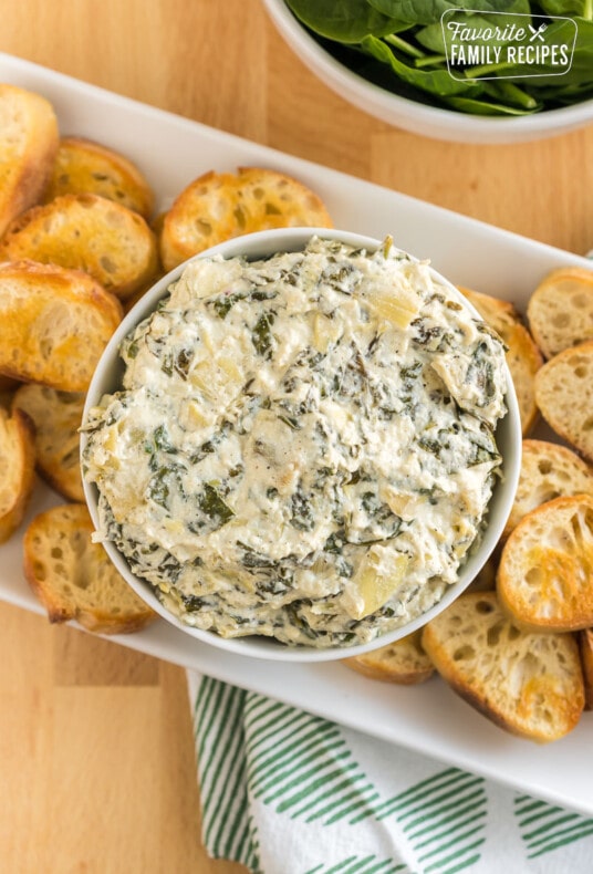 A bowl of crock pot spinach artichoke dip surrounded by slices of baguette