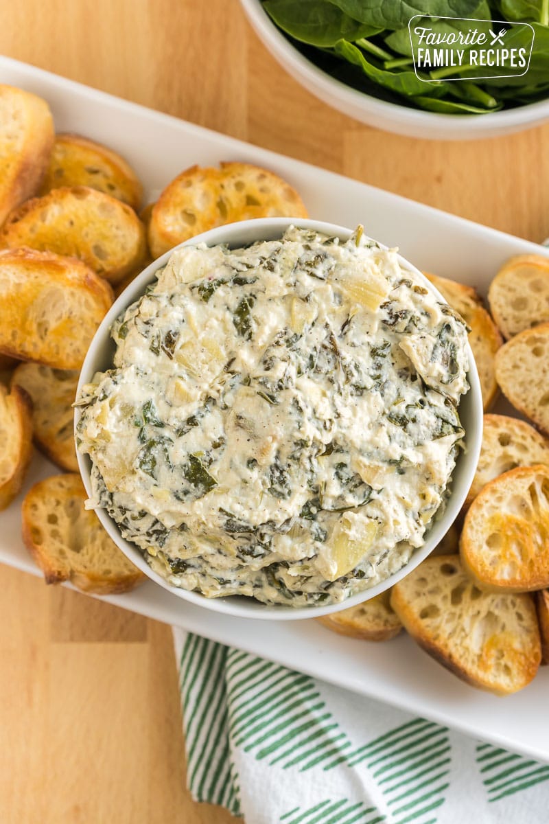 A bowl of crock pot spinach artichoke dip surrounded by slices of baguette.