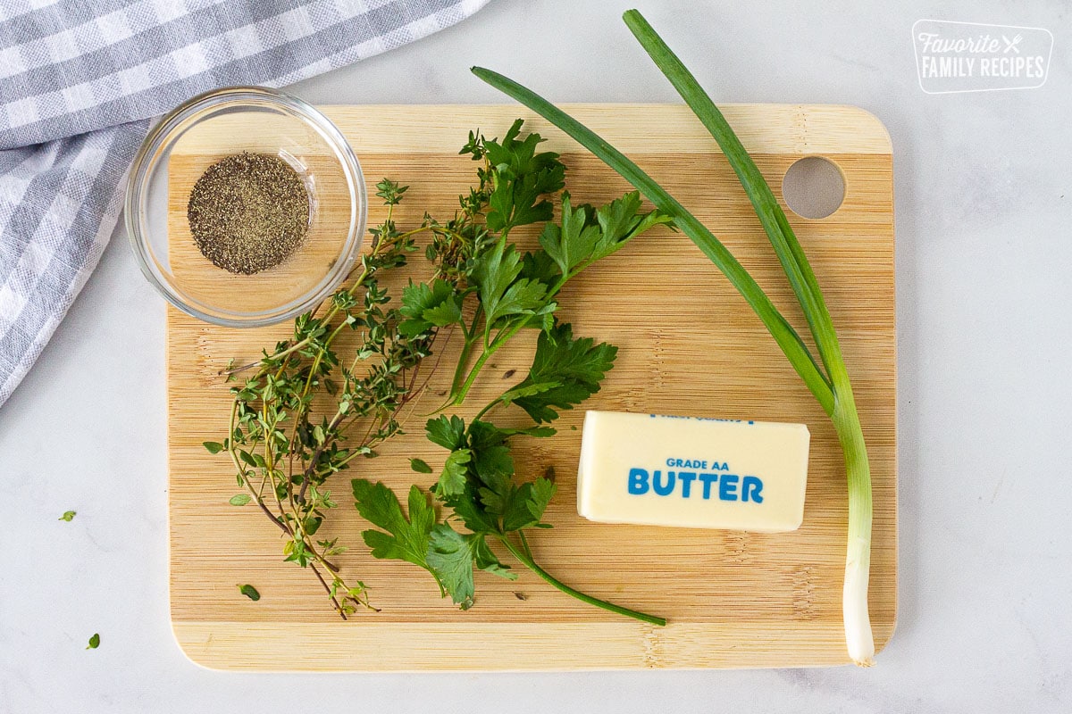 Cutting board with thyme, parsley, green onion, butter and pepper in a bowl to make herbed butter.