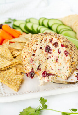 Cranberry Almond Bacon Cheese Ball missing a wedge. Crackers and vegetables surrounding the cheese ball.