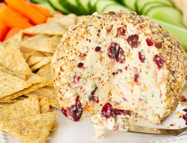 Cranberry Almond Bacon Cheese Ball missing a wedge. Crackers and vegetables surrounding the cheese ball.