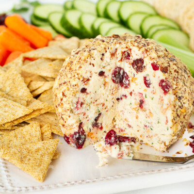 Cranberry Almond Bacon Cheese Ball missing a wedge. Crackers and vegetables surrounding the cheese ball.