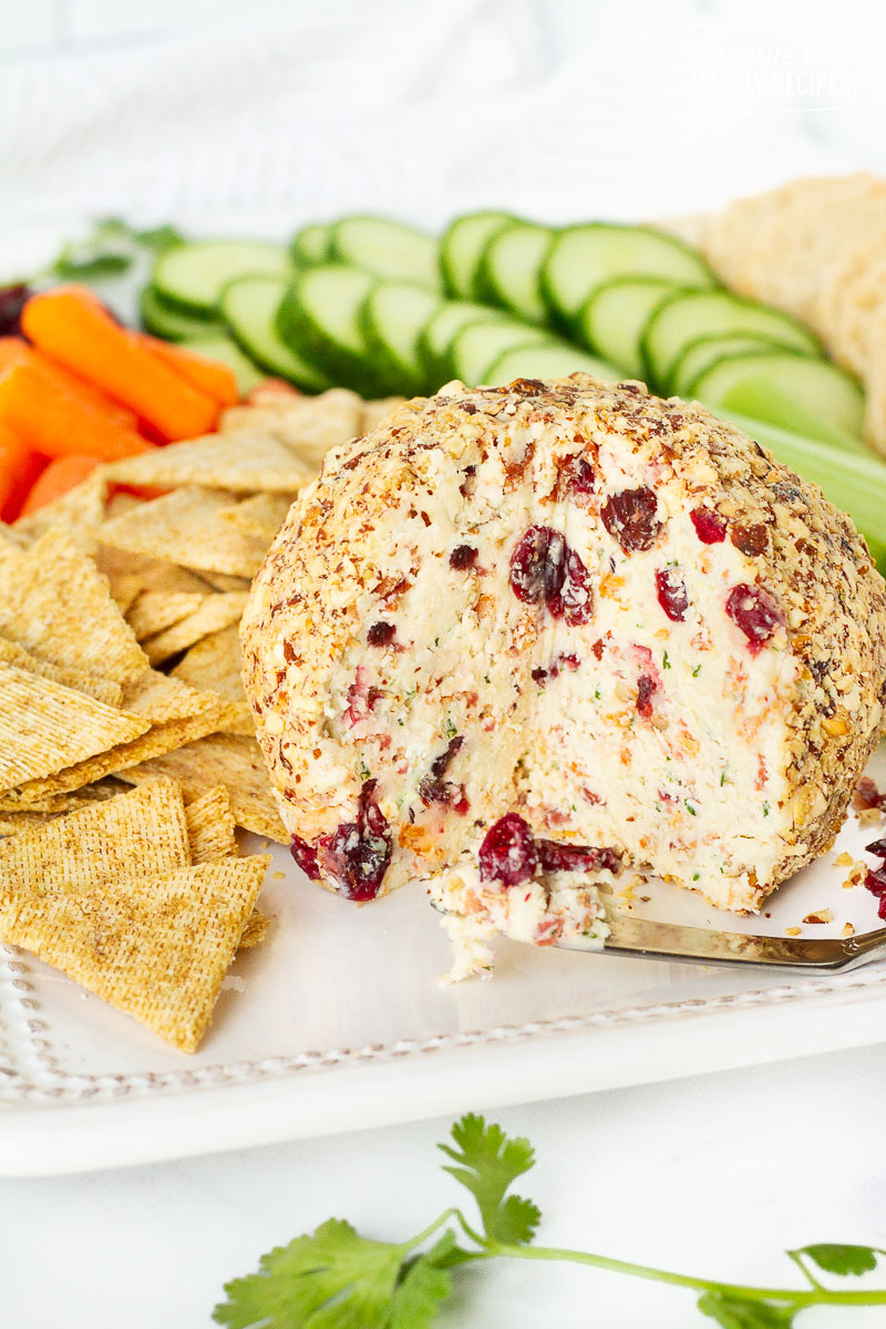 Cranberry Almond Bacon Cheese Ball missing a wedge. Crackers and vegetables surrounding the cheese ball.