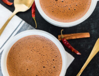 Two mugs of Mexican hot chocolate on a tabletop with cinnamon sticks and chiles