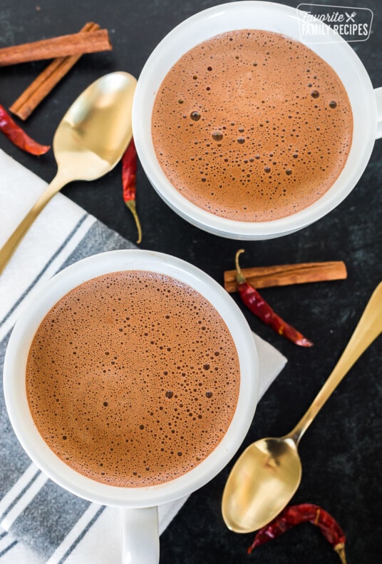 Two mugs of Mexican hot chocolate on a tabletop with cinnamon sticks and chiles