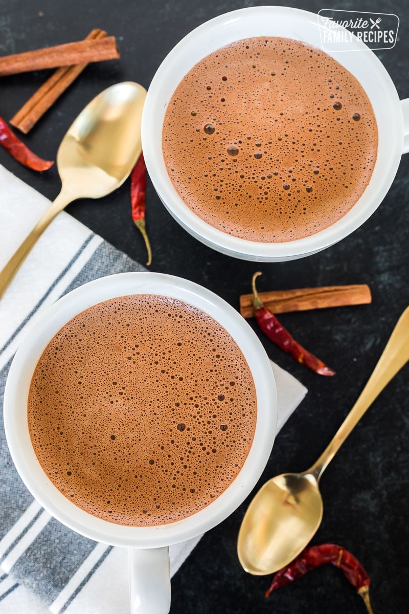 Two mugs of Mexican hot chocolate on a tabletop with cinnamon sticks and chiles.