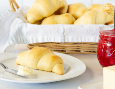 Plate with a Homemade Crescent Roll and butter with a baked of rolls behind. Jam and butter on the side.