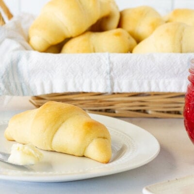 Plate with a Homemade Crescent Roll and butter with a baked of rolls behind. Jam and butter on the side.