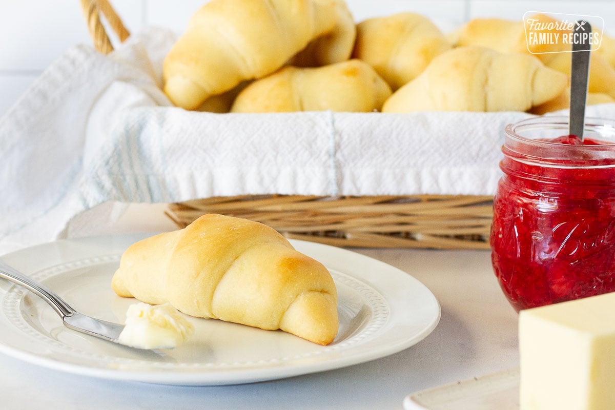 Plate with a Homemade Crescent Roll and butter with a baked of rolls behind. Jam and butter on the side.