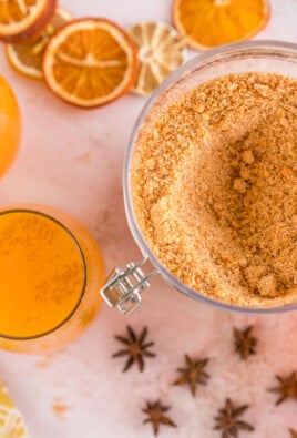 A large container of dry wassail mix next to some glasses full of wassail recipe