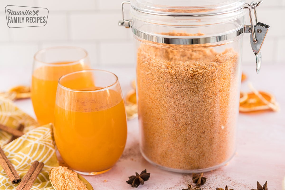 A large container of dry wassail mix next to some glasses full of wassail.