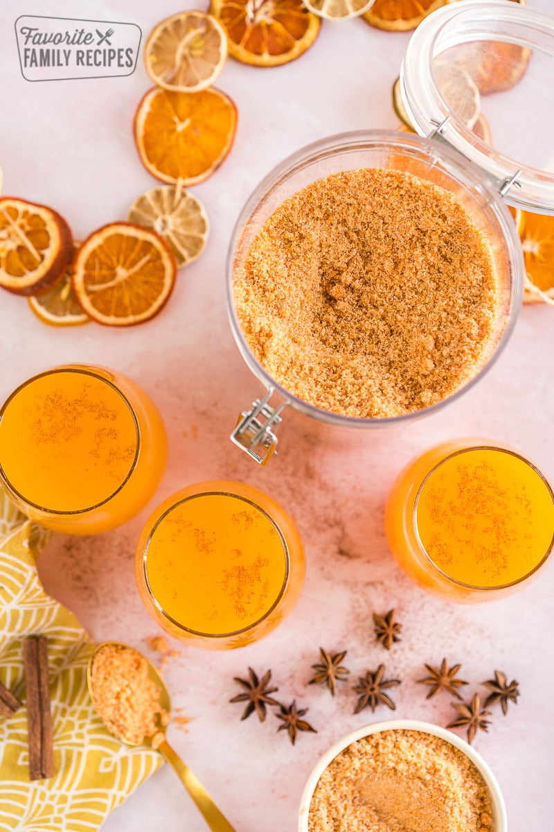 A large container of dry wassail mix next to some glasses full of wassail recipe.