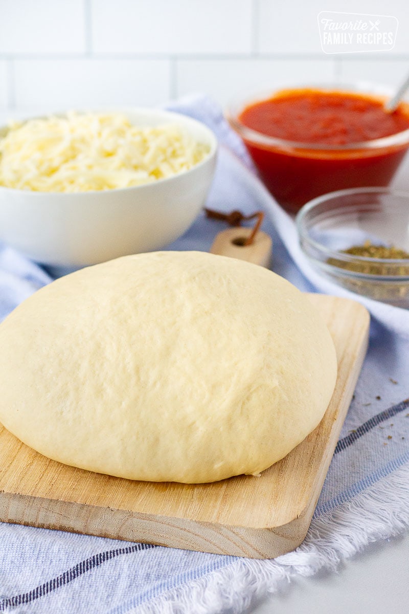 Homemade Pizza Dough shaped in a ball on a cutting board. Cheese, sauce, a oregano on the side.