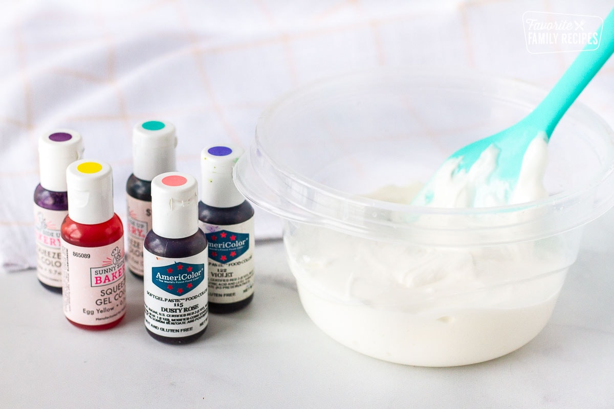 Gel paste colors next to a plastic mixing bowl of royal icing with spatula.