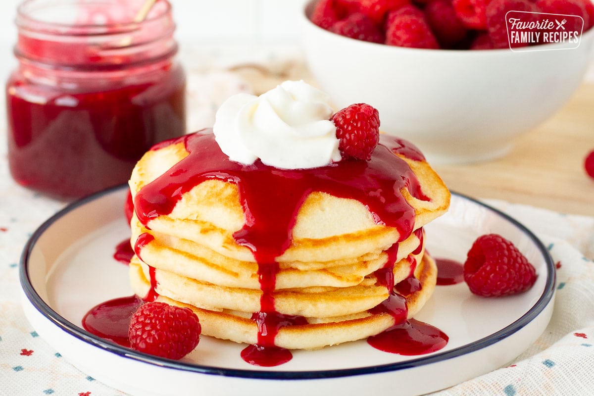 Stack of pancakes with raspberry coulis, fresh raspberries and whipped cream on a plate.