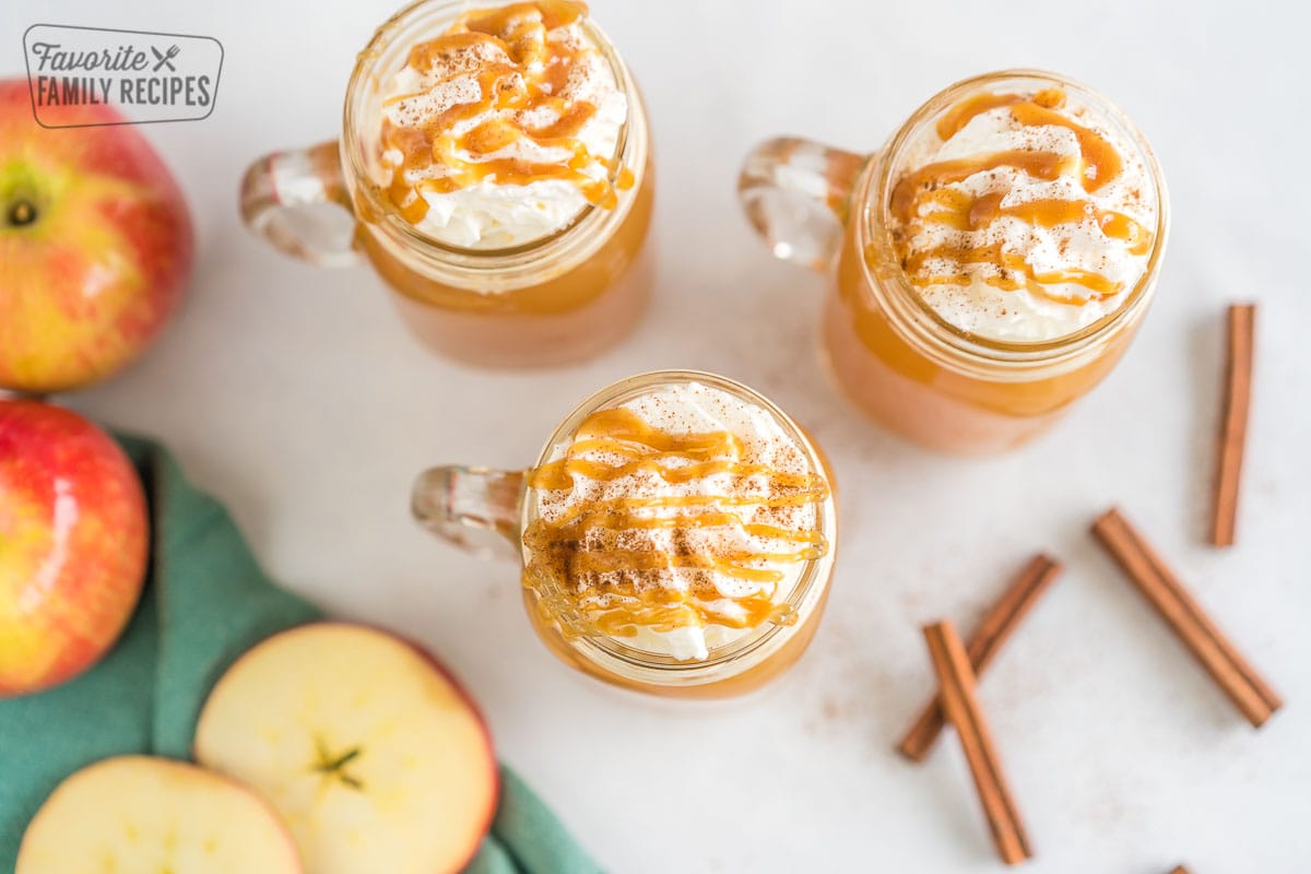 Three mugs of Starbucks Caramel Apple Spice Cider topped with whipped cream and caramel