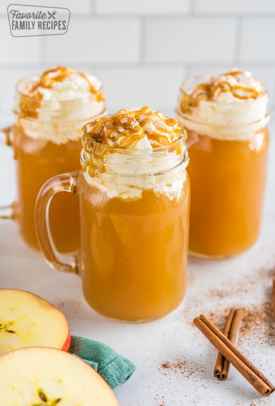 Three mugs of Starbucks Caramel Apple Spice Cider topped with whipped cream and caramel