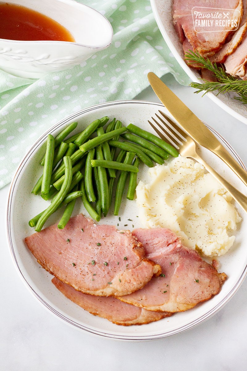 Ham, mashed potatoes, and green beans on a white plate with gold silverware. 