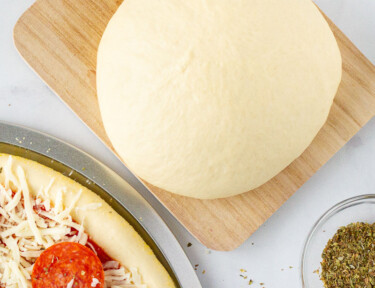 Ball of Homemade Pizza Dough on a board next to an unbaked pepperoni pizza.