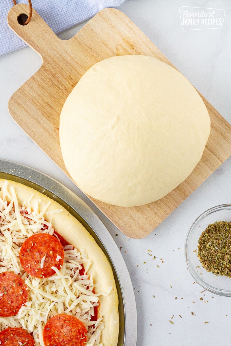 Ball of Homemade Pizza Dough on a board next to an unbaked pepperoni pizza.