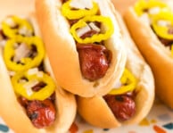 Four hot dogs in buns on a plate topped with onions and banana peppers
