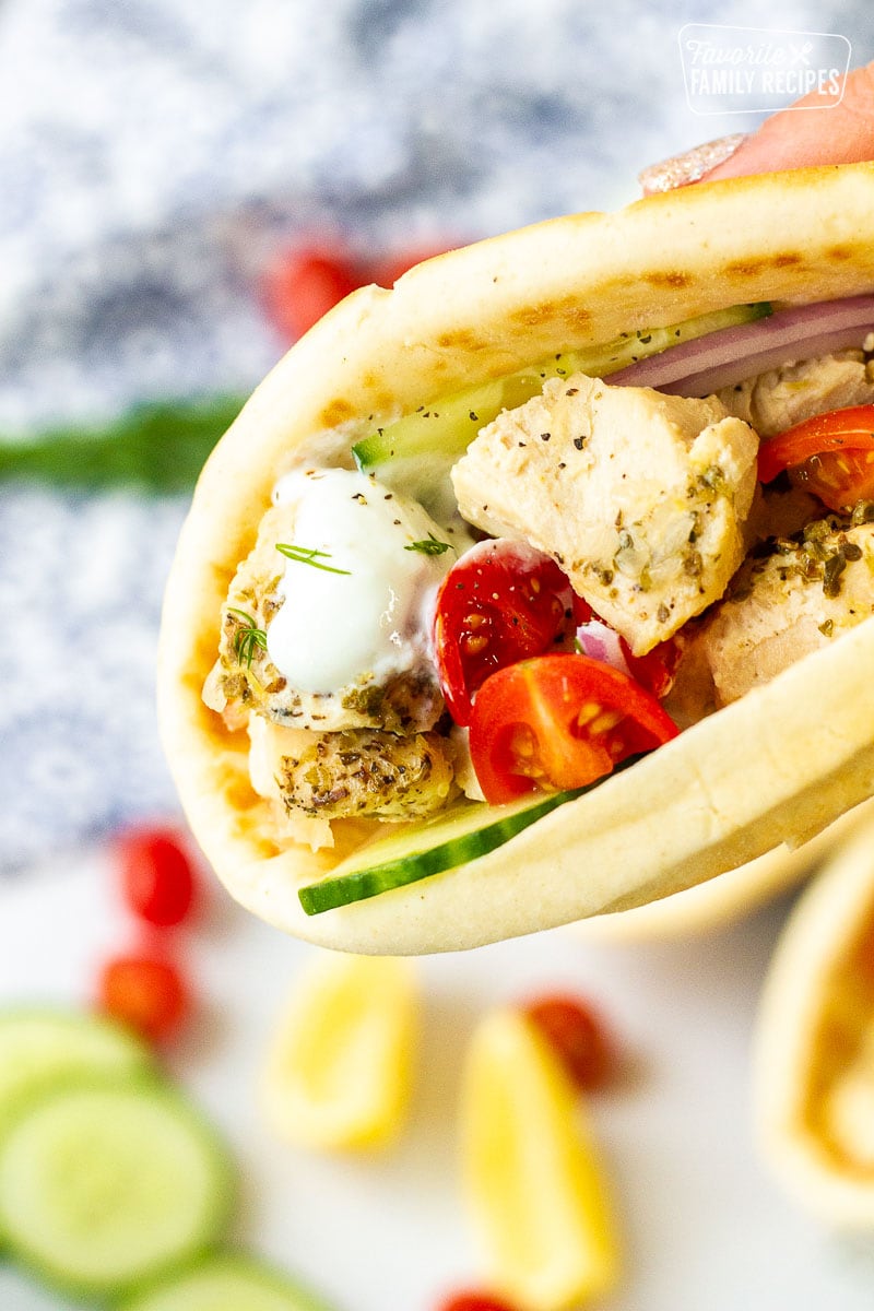 Hand holding a Chicken Gyro with tomatoes, cucumbers, tzatziki sauce and red onions.