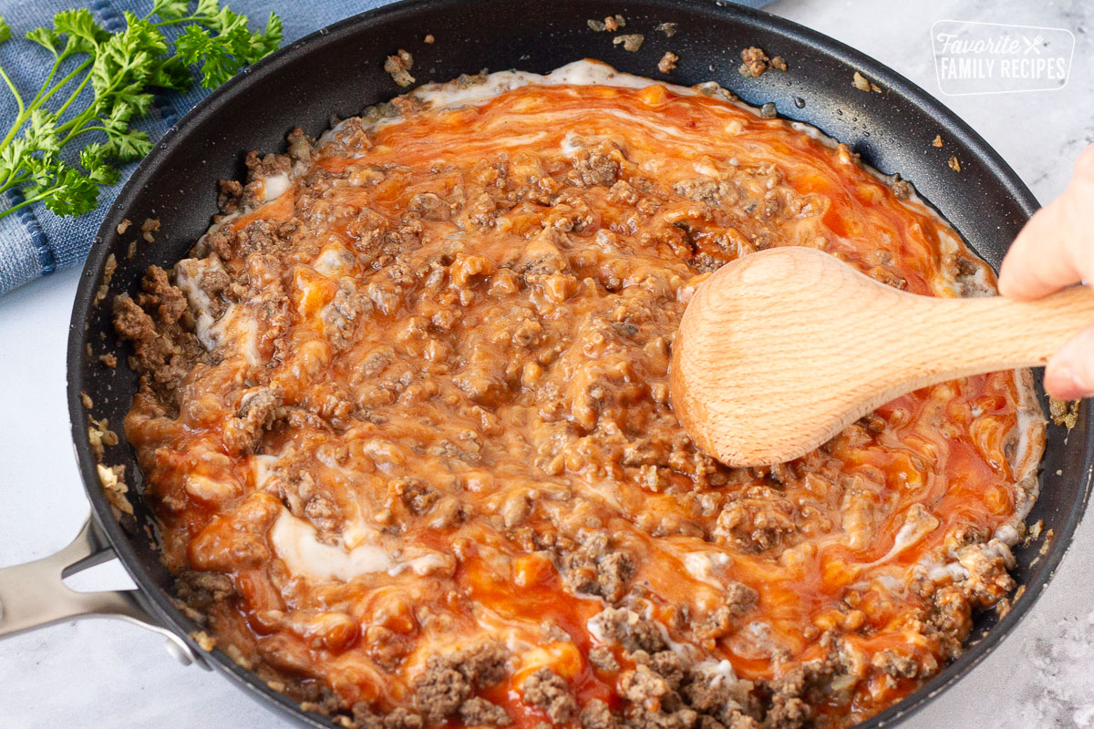 Wooden spoon mixing ground beef and sauces together in a skillet for Creamy Ground Beef and Noodles.