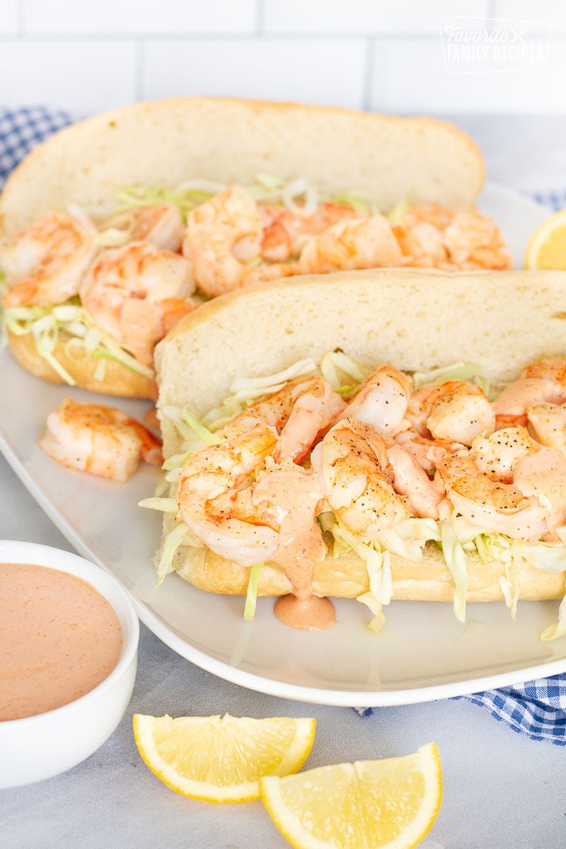 Two Shrimp Po'Boy Sandwiches on a plate with cajun sauce and lemon wedges.