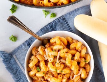 Small bowl of Creamy Ground Beef and Noodles Skillet with a fork next to a skillet of Creamy Ground Beef and Noodles. Bread sticks on the side.